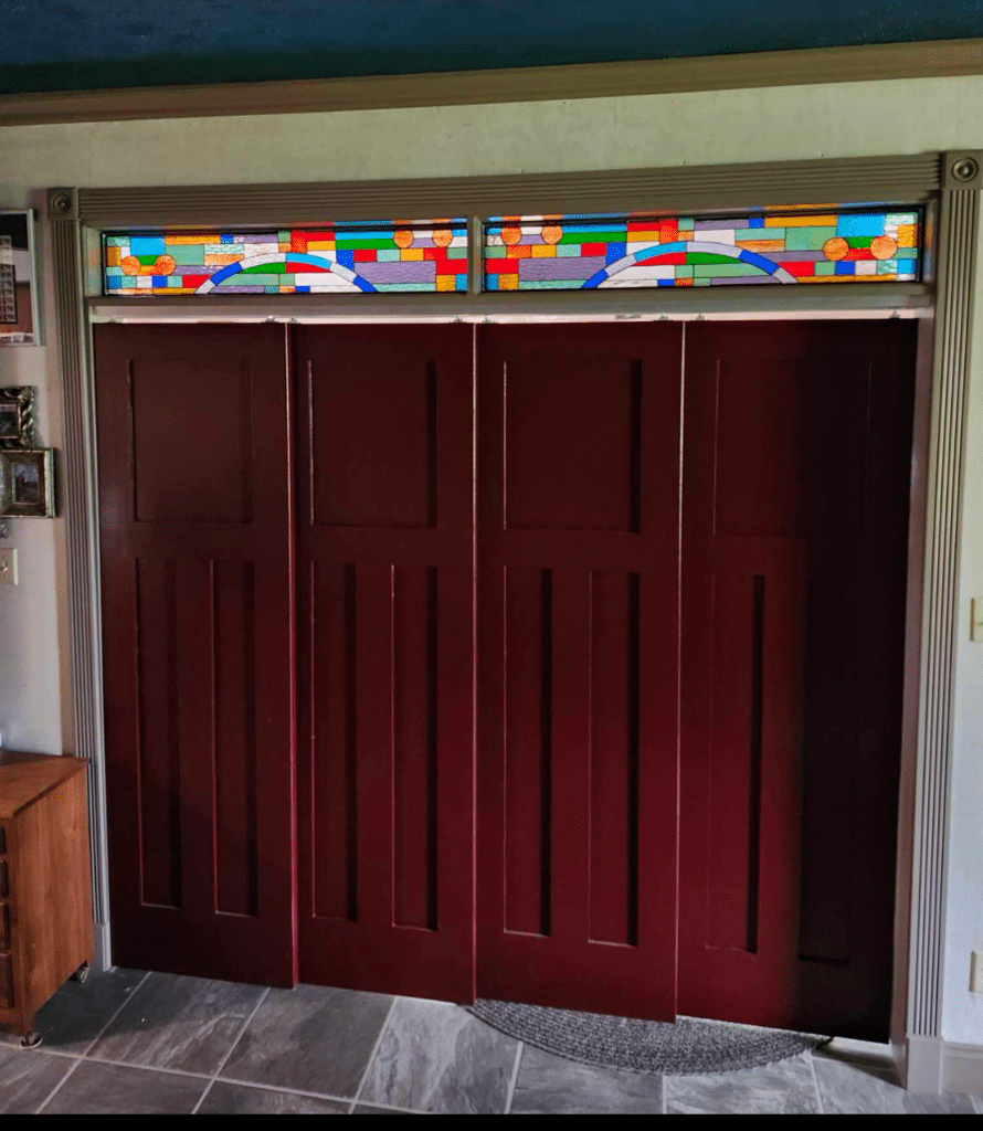 A rectangular stained glass transom window with a vibrant, abstract geometric design featuring an arch and circular elements, installed above four red bifold doors.